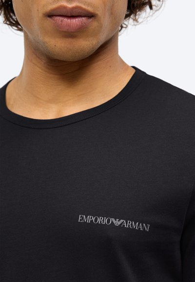 T-shirt de algodão preto com decote redondo. Apresenta um logo em prata no lado esquerdo do peito. Textura suave e design ajustado.