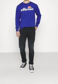 Sweatshirt bleu à manches longues avec le logo « ellesse » en blanc et orange, assorti à un jean noir et des baskets montantes noir/blanc.