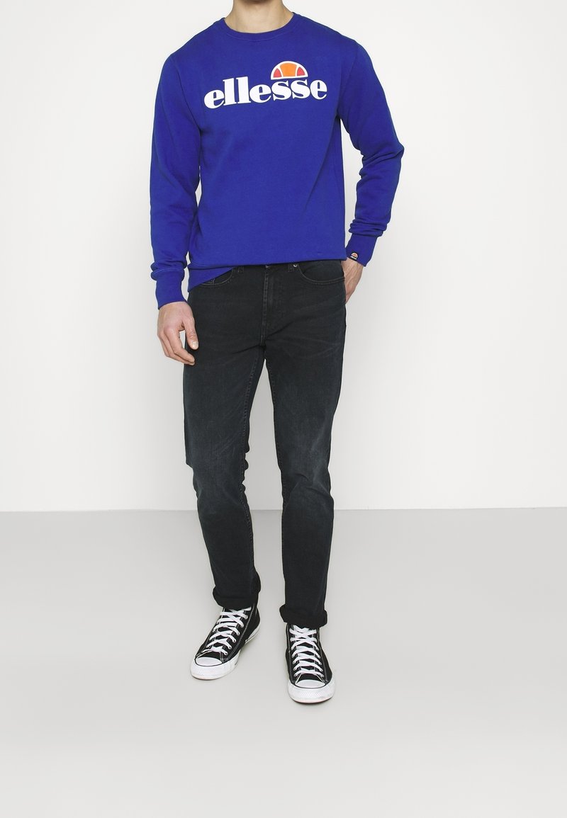 Sweatshirt bleu à manches longues avec le logo « ellesse » en blanc et orange, assorti à un jean noir et des baskets montantes noir/blanc.