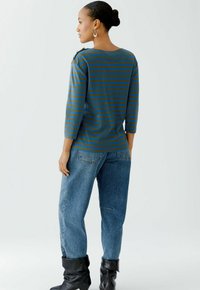Gestreiftes Strickoberteil in Blau und Oliv mit Dreiviertelärmeln und Schulterdetails; kombiniert mit lockeren Jeans und schwarzen Stiefeletten.