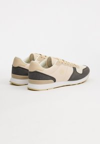 Zapatillas beige y grises con un upper de tela texturizada, acentos sintéticos suaves y una suela de goma blanca, que presentan detalles de marca en tonalidades.