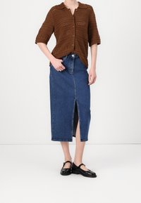 Suéter marrón de punto con botones y mangas cortas, combinado con una falda vaquera azul con una abertura frontal. El conjunto se completa con zapatos negros.