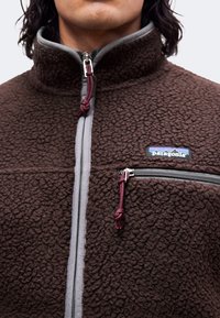 Κοντινή λήψη στήθους ατόμου που φοράει καφέ fleece μπουφάν Patagonia με γκρι φερμουάρ και θήκη στήθους με φερμουάρ και σκούρες κόκκινες κορδόνια.