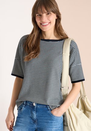 Junge Frau lächelnd, trägt ein schwarz-weiß gemustertes kurzärmeliges Oberteil, blaue Jeans und eine beige Schultertasche, eine Hand in der Tasche.