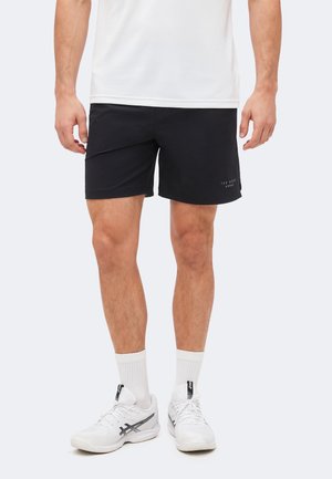 Férfi fekete sportnadrágban, fehér bokazokniban és fehér Asics tornacipőben áll, karjai nyugalmi helyzetben oldalán.