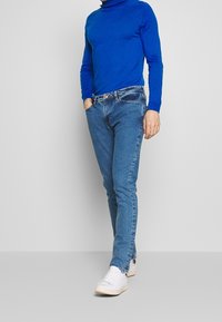 Blå polotröja med långa ärmar i kombination med ljusblå, slim-fit jeans och vita sneakers. Påtaglig sömnad och detaljer i hårdvara.