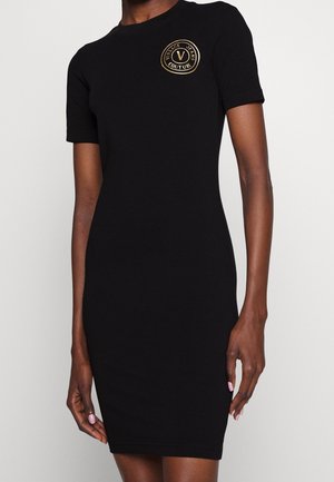 Schwarzes, kurzärmeliges, figurbetontes Kleid mit goldenem rundem Logo mit der Aufschrift "Versace Jeans Couture" auf der oberen linken Brust.