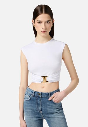Top bianco corto con maniche a cappuccio, caratterizzato da una fibbia decorativa dorata sul davanti e una texture liscia, abbinato a jeans in denim blu.