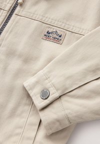 bézs dzseki texturált anyagból, cipzáras záródással, logóval ellátott folttal és fém patentgombbal az ujjatlanon.