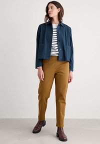 Giacca cropped blu navy con chiusura a bottoni e tasche frontali indossata sopra una camicia a righe bianche e blu navy, abbinata a pantaloni a gamba affusolata marroni e stivaletti.