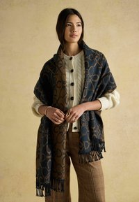 Modrý a hnedý vzorovaný šál s strapcami prehodený cez béžový sveter, obsahujúci textúrovaný pletený dizajn a prispôsobené kockované nohavice.