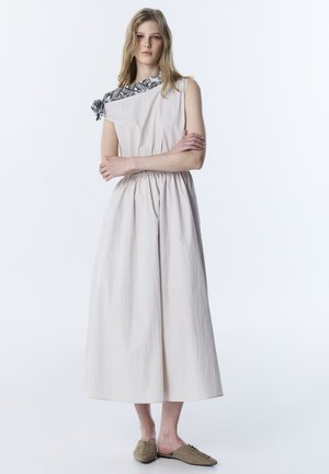 Mujer vestida con un vestido midi blanco sin mangas, con detalle en los hombros en blanco y negro, y zapatos beige sin cordones, de pie con los brazos cruzados.