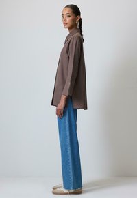 Camisa marrón de manga larga con cuello, combinada con vaqueros de talle alto en azul claro. Los zapatos son zapatillas beige. Diseño simple y moderno.