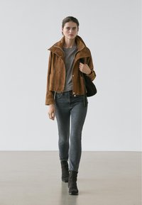 Jeune femme marchant, portant une veste en daim marron, un t-shirt gris, un jean noir délavé, des bottines noires, et portant un sac à bandoulière noir.