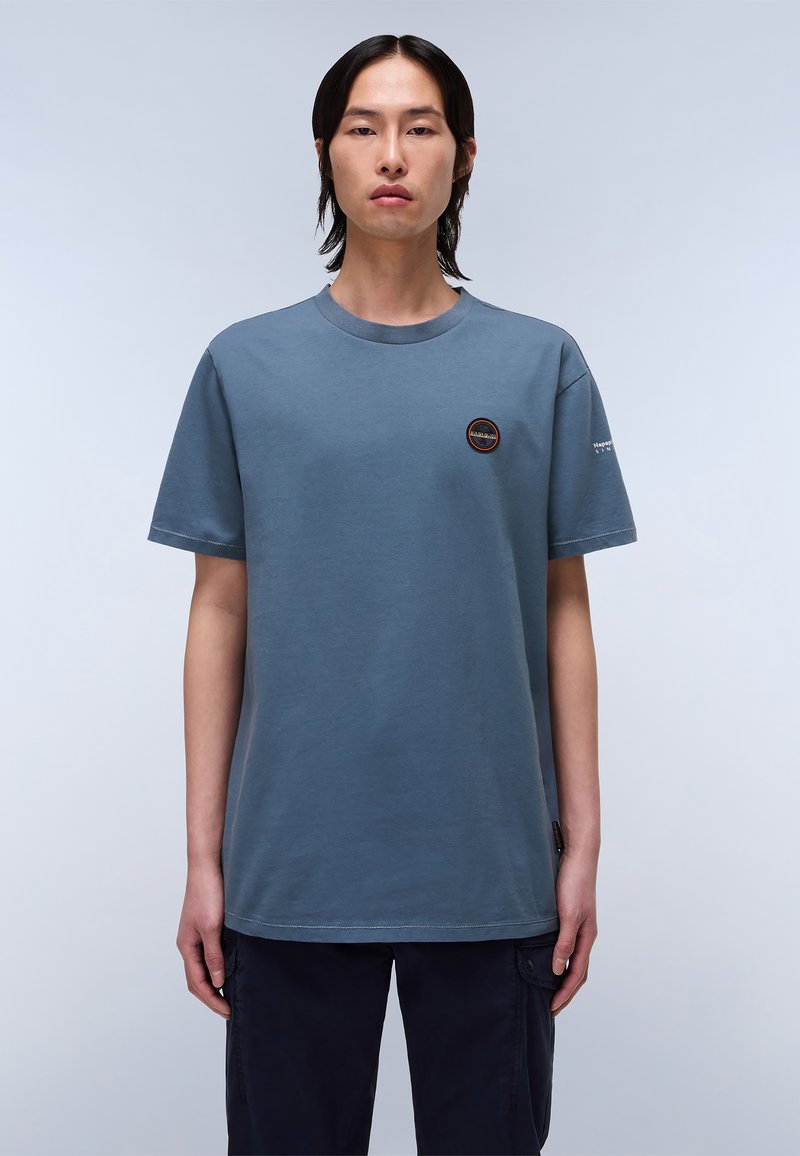 Camiseta de algodón azul con cuello redondo, manga corta y un pequeño logo circular en el pecho. Presenta un corte relajado y una textura suave.