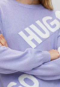Lichtpaarse gebreide trui met witte tekst "HUGS"; heeft geribde halslijn en omgeslagen mouwen; textuurstof.
