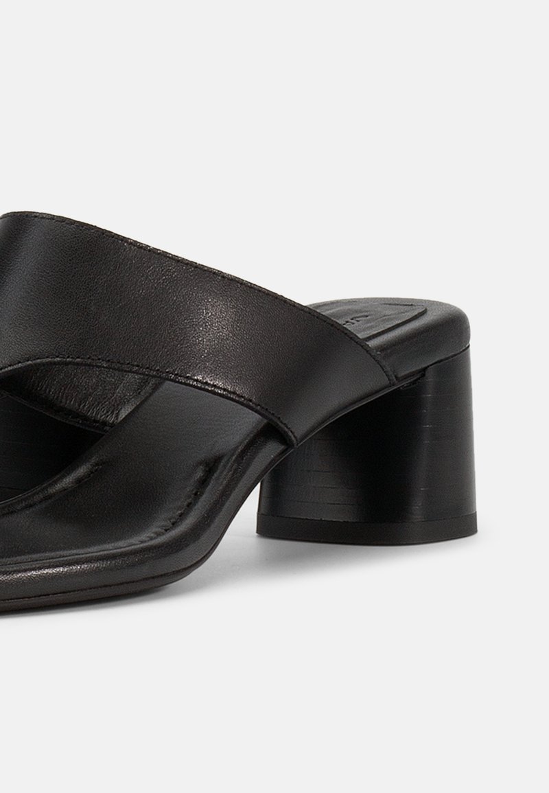 Schwarze Leder-Mulesandale mit breiten, gekreuzten Riemen und kurzem, klobigem Blockabsatz vor weißem Hintergrund.