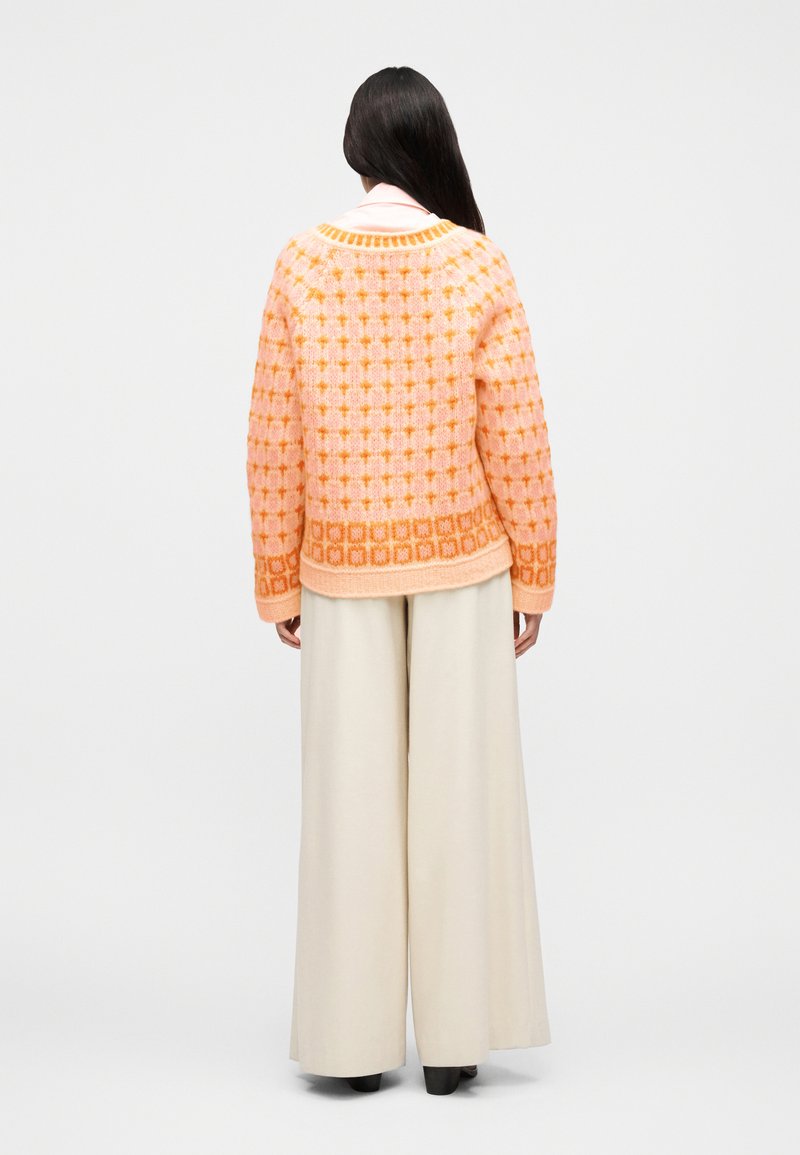 Femme aux longs cheveux foncés vue de dos, portant un pull à motifs roses et orange, un pantalon large crème et des chaussures noires.