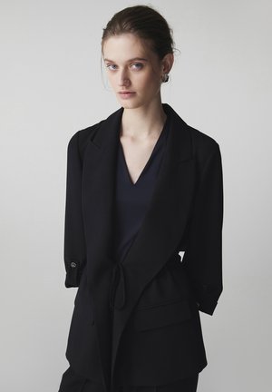 Jeune femme à la peau claire et aux yeux bleus, portant un blazer noir aux manches retroussées et un haut sombre à encolure en V, posant devant un fond uni.