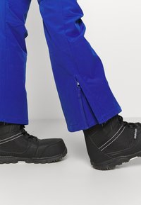 Niebieskie wodoodporne spodnie z zamkami błyskawicznymi po bokach, w zestawie z czarnymi ocieplanymi butami z białymi szwami i gumowymi podeszwami.