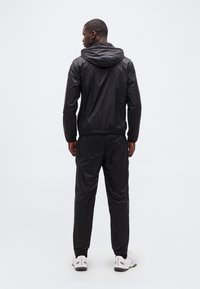 Άνδρας με την πλάτη γυρισμένη, φοράει μα ύρο μπουφάν με κουκούλα, μαύρο παντελόνι που ταιριάζει και λευκά αθλητικά παπούτσια, με φόντο ένα απλό φωτεινό τοπίο.