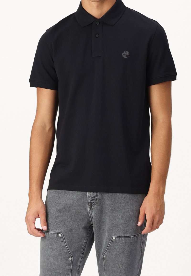 Homme portant un polo noir à manches courtes avec un petit logo d'arbre sur la poitrine et un jean gris avec des poches avant.