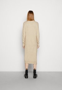 Beige pletené šaty s dlouhými rukávy a žebrovaným lemem, s volným střihem. Nošeny s černými kotníkovými botami, na jednobarevném pozadí.