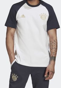 Bílé tričko s krátkým rukávem a tmavě modrými raglánovými rukávy, s logem Adidas ve zlaté barvě a emblémem FC Bayern München na hrudi. Směsová tkanina z bavlny.