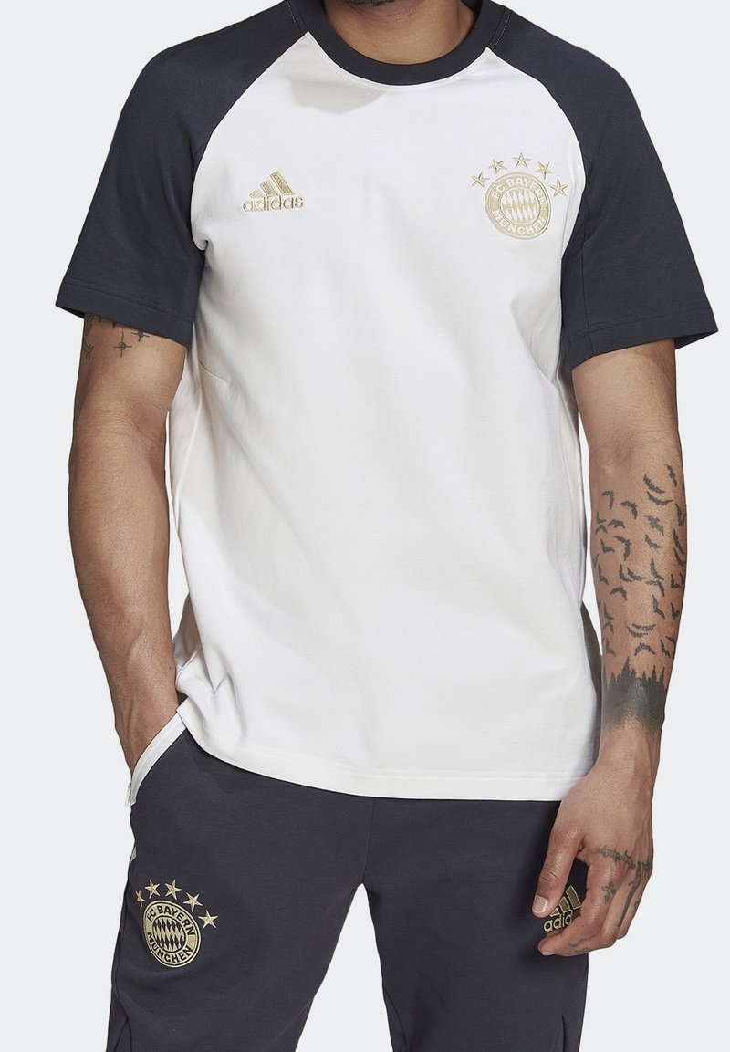 Bílé tričko s krátkým rukávem a tmavě modrými raglánovými rukávy, s logem Adidas ve zlaté barvě a emblémem FC Bayern München na hrudi. Směsová tkanina z bavlny.