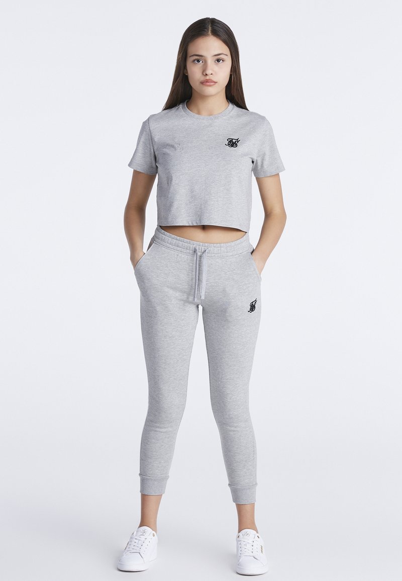 Camiseta gris de corte corto con mangas cortas, con un logo negro; combinada con pantalones elásticos grises a juego y zapatillas blancas.