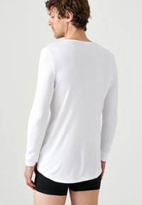 Witte shirt met lange mouwen en ribbelstof, gemaakt van elastische stof. Heeft een ronde hals en een afgeronde zoom, gedragen over zwarte shorts.