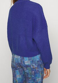 Lila stickad tröja med strukturerat mönster och ribbad nederkant, tillsammans med blå mönstrade byxor med en grafisk design av ett digitalt öga.