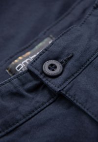 Gros plan sur un tissu bleu foncé avec un bouton noir marqué "Ombre Wear" et des coutures visibles près d'une couture pliée.