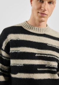 Czarny i kremowy sweter w paski o teksturowanym wykończeniu, z prążkowanym dekoltem i nierównymi wzorami pasków na całej powierzchni.
