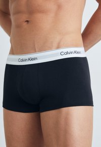 Crne bokserice od pamuka s bijelo-sivim elastičnim pojasom na kojem se nalazi natpis "Calvin Klein". Glatka tekstura, uski kroj.