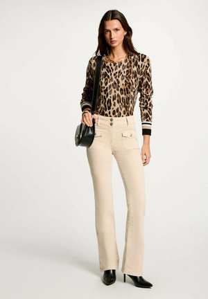 Pantaloni beige a zampa d'elefante con tasche frontali, abbinati a un maglione marrone con stampa leopardata. La modella porta una borsa a tracolla nera e indossa tacchi neri.