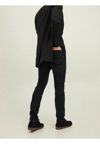 Černý blazer přes tmavý svetr, doplněný o slim černé kalhoty a černé semišové boty se světle hnědou podrážkou. Oblečení je monochromatické.
