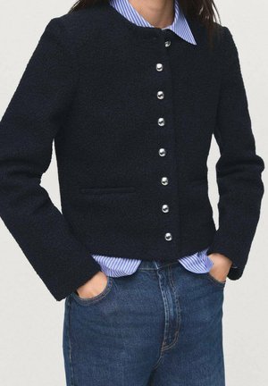 Giacca testurizzata blu navy con bottoni argentati, scollo rotondo e tasche laterali, indossata sopra una camicia a righe blu chiaro.
