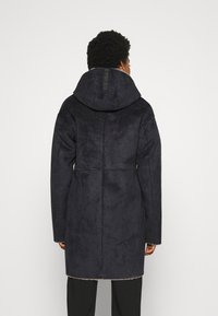 Cappotto blu navy scuro realizzato in un morbido tessuto texturizzato; presenta un ampio cappuccio e cuciture bianche lungo le giunture. La vista posteriore mostra un design pulito e slanciato.
