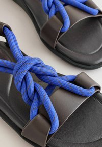 Sandales noires avec larges brides marron et lacets épais en corde bleue noués en nœuds décoratifs sur le dessus.