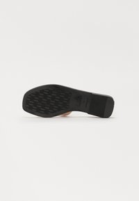 Semelle en caoutchouc noir avec un motif de texture antidérapant et un léger talon, attachée à un matériau supérieur de couleur beige clair. Aucun détail de marque visible.