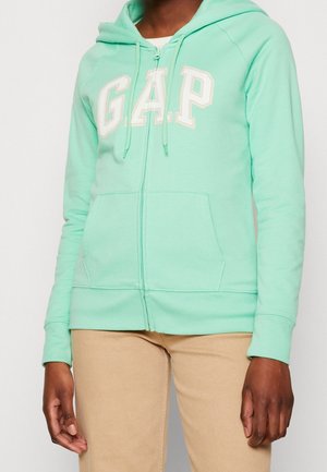Person wearing a mint green GAP zip-up hoodie with front pockets and beige pants, standing against a plain background.