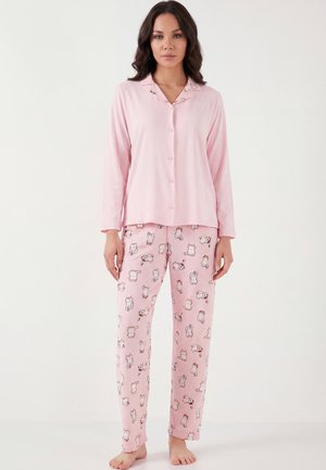 Femeie în picioarele goale, purtând un top de pijama roz deschis cu nasturi și pantaloni asortați, cu mici ilustrații de pisici, pe un fundal alb simplu.