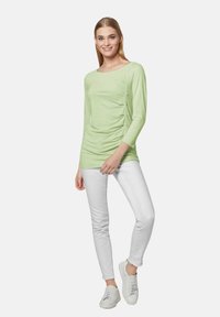 Hellgrünes Langarm-Oberteil mit seitlichen Raffungselementen, kombiniert mit weißen schmalen Jeans und weißen Sneakern. Glatte Stoffoberfläche.