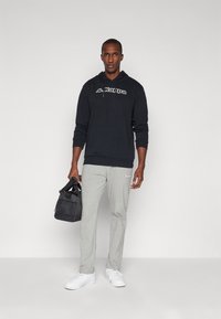 Felpa nera con cappuccio e logo, pantaloni della tuta grigi e scarpe da ginnastica bianche. Porta una piccola borsa nera. Abbigliamento sportivo semplice e casual.