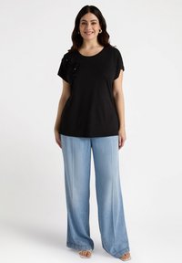 Czarna koszulka z krótkim rękawem z teksturowanymi zdobieniami na lewym ramieniu, w połączeniu z luźnymi jasnoniebieskimi spodniami dżinsowymi.