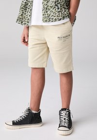 Beige shorts med sidfickor och broderad branding. Texturerat, lätt tyg. Svarta höga sneakers med vita skosnören och blixtlås.
