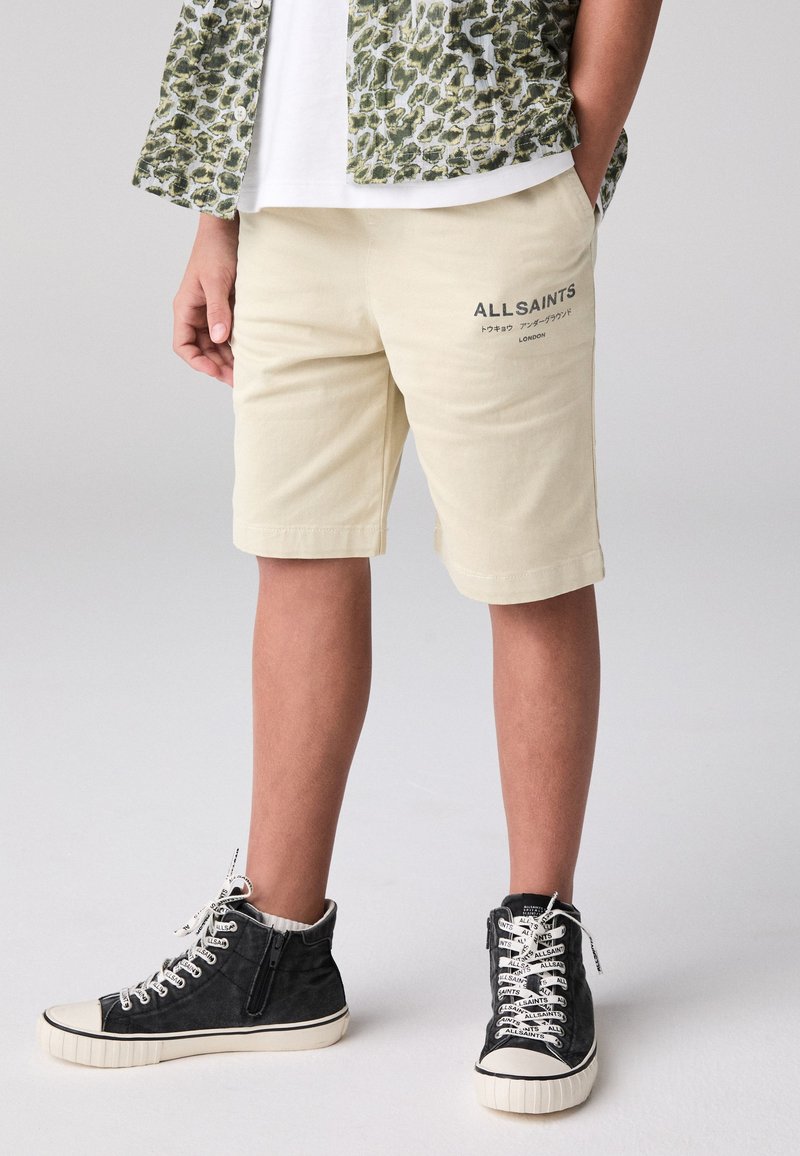 Beige shorts med sidfickor och broderad branding. Texturerat, lätt tyg. Svarta höga sneakers med vita skosnören och blixtlås.
