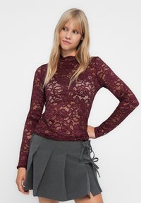 Ung kvinne iført en gjennomsiktig burgunder blomstermønstret bluse i blonde og grå pressede skjørt med sideløkker, stående mot en enkel hvit bakgrunn.