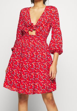 Rode bloemenjurk met lange opgeblazen mouwen, knoopsluiting aan de voorkant en een buikuitsparing, gedragen door een staand persoon tegen een effen achtergrond.